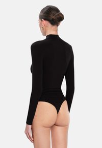 Femme avec les cheveux en chignon portant un bodysuit à manches longues noir avec col roulé, vue de dos sur un fond clair et uni.