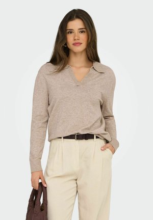Mujer con cabello largo castaño, vestida con un suéter beige de cuello en V y pantalones crema claros, sosteniendo un bolso marrón oscuro con una mano, y la otra mano en el bolsillo.