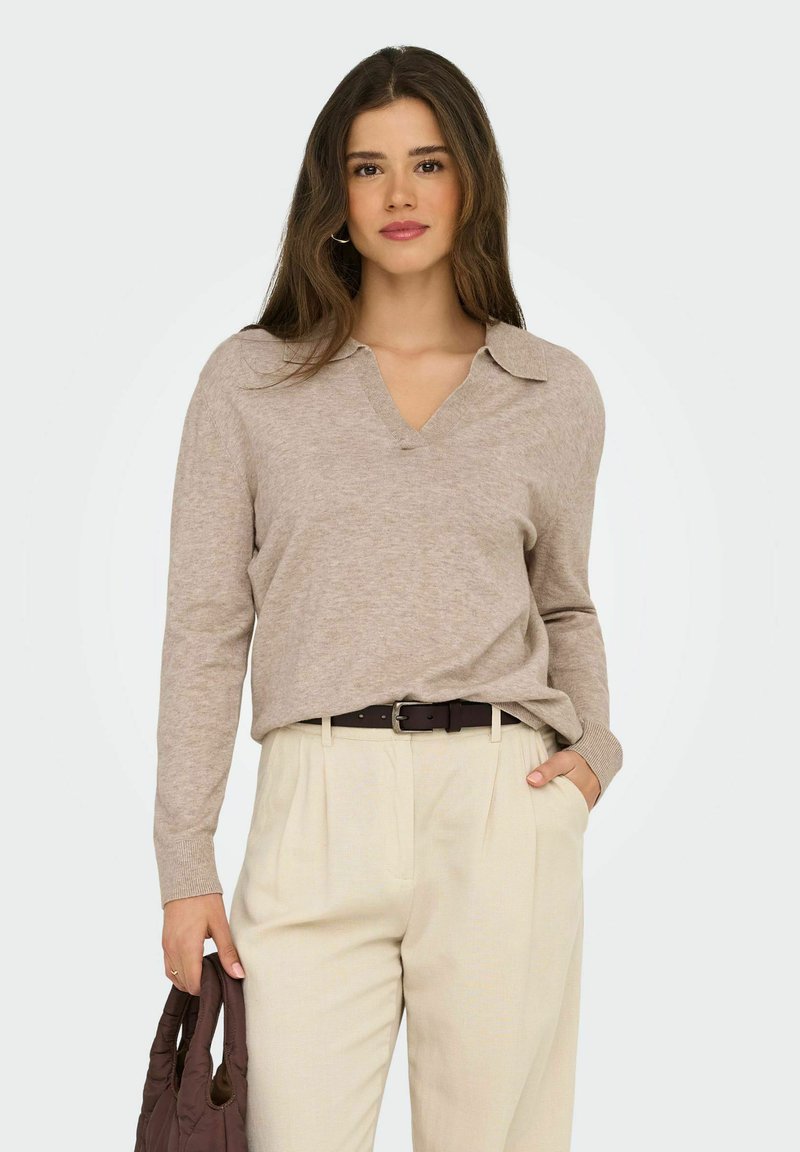 Mujer con cabello largo castaño, vestida con un suéter beige de cuello en V y pantalones crema claros, sosteniendo un bolso marrón oscuro con una mano, y la otra mano en el bolsillo.