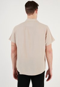 Camisa de manga corta en tela beige clara, con un corte holgado, costura en el canesú trasero y dobladillo redondeado. Presenta una textura horizontal sutil.