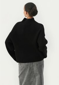 Schwarzer gerippter Strickpullover mit hohem Kragen, gesenkten Schultern und lockerer Passform, kombiniert mit einem grauen Jeansrock. Der strukturierte Materialdetail ist sichtbar.