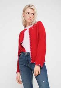 Κόκκινη πλεκτή ζακέτα με υφή, που διαθέτει ξεφτισμένο τελείωμα, ροζ λεπτομέρειες και κουμπιά μπροστά, φορεμένη πάνω από ανοιχτό ροζ τοπ.