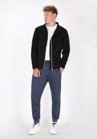 Giacca zip-up nera con accenti bianchi, abbinata a pantaloni jogger blu navy con tasche laterali e toppe sulle ginocchia. Il modello indossa sneaker bianche.