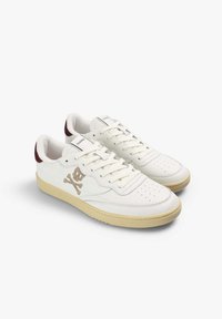 Zapatillas de cuero blanco con un diseño retro, que presentan un logotipo de calavera y huesos cruzados, puntera perforada y suela de goma beige. Acento en el talón en color burdeos.