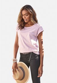 Rosa kurzärmliges Oberteil mit strukturiertem Muster, kombiniert mit schwarzen Hosen. Das Model hält einen Strohhut mit bunten Bandakzenten.