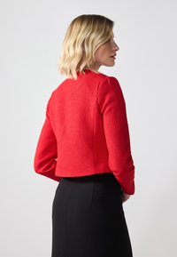 Veste courte texturée rouge à manches longues et col rond, associée à une jupe noire, vue de dos.