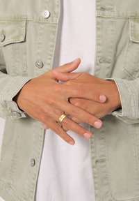 Anello d'oro su dita, mani unite. Giacca di jeans verde chiaro con bottoni d'argento sopra una camicia bianca. Texture della pelle liscia visibile.