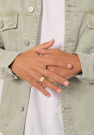 Goldene Ringe an den Fingern, Hände gefaltet. Hellgrüne Jeansjacke mit silbernen Knöpfen über einem weißen Hemd. Glatte Hautstruktur sichtbar.