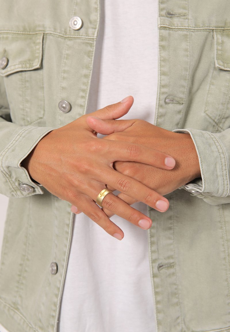 Anello d'oro su dita, mani unite. Giacca di jeans verde chiaro con bottoni d'argento sopra una camicia bianca. Texture della pelle liscia visibile.