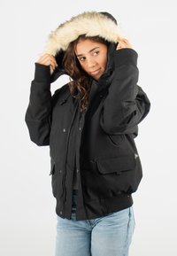 Veste noire avec capuche en fourrure, dotée d'une fermeture éclair, de deux grandes poches avant et de poignets côtelés. Texture lisse et coupe ample.