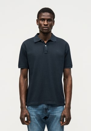 Άνδρας που φοράει μαύρο πόλο με κοντά μανίκια και μπλε τζιν, στέκεται με το πρόσωπο προς τα εμπρός σε απλό ανοιχτόχρωμο φόντο.