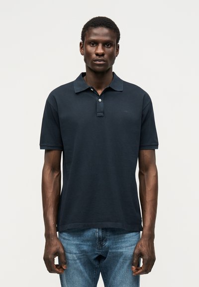 Homme portant un polo noir à manches courtes et un jean bleu, debout face à un fond clair uni.