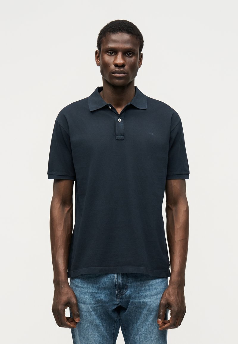 Άνδρας που φοράει μαύρο πόλο με κοντά μανίκια και μπλε τζιν, στέκεται με το πρόσωπο προς τα εμπρός σε απλό ανοιχτόχρωμο φόντο.