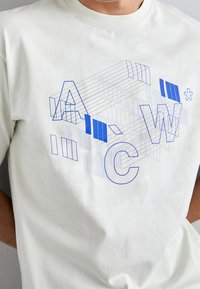 Krátký rukáv, světle krémové tričko s modrou grafikou a stylizovanými písmeny a geometrickými liniemi, vyrobené z bavlny.