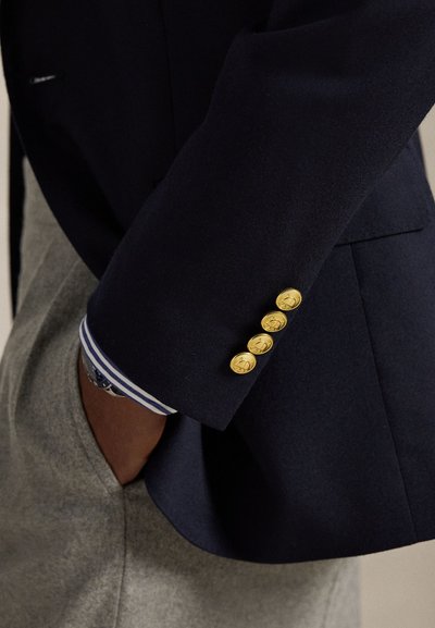 Blazer azul marinho com botões dourados na manga, apresentando punhos às riscas azuis e brancas. Tecido cinzento visível a partir da cintura, de textura suave.