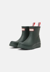 Botas de goma oscuras verdes hasta el tobillo con puntera redondeada, suela texturizada y tiradores a rayas rojas y blancas; presenta el logo "HUNTER" en la parte frontal.