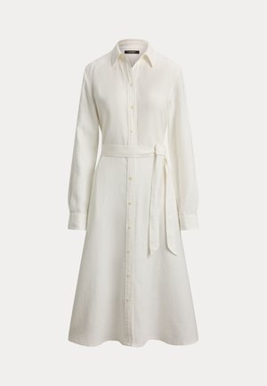 Robe chemise blanche à manches longues avec boutons, col et ceinture à nouer à la taille, jupe trapèze sur un fond uni.
