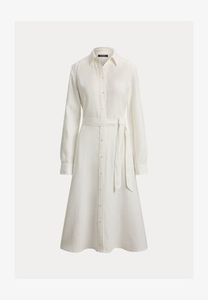 Robe chemise blanche à manches longues avec boutons, col et ceinture à nouer à la taille, jupe trapèze sur un fond uni.