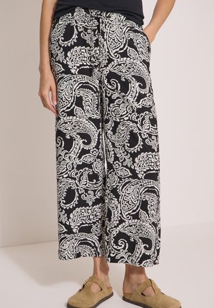 Pantalon noir à jambes larges avec motif cachemire blanc porté avec des chaussures en daim beige sans lacets et un haut noir partiellement visible.