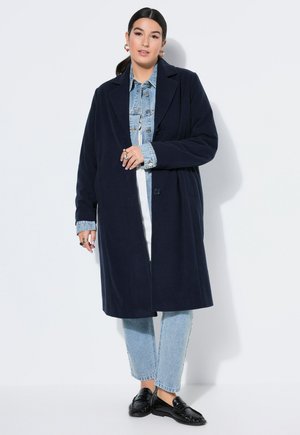 Kvinde iført en lang marineblå frakke over en lyseblå cowboyjakke og jeans, stående med den ene hånd, der holder frakkens forside.