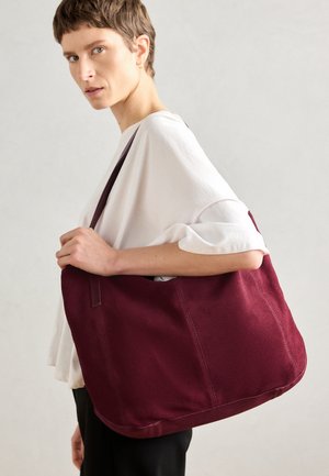 Personne portant un haut blanc et un pantalon noir tient un grand sac à épaule bordeaux sur un fond clair uni.