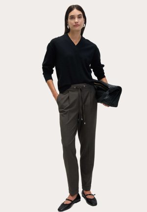 Kobieta ubrana w czarny sweter z dekoltem w serek, szare spodnie z wysokim stanem i sznurkiem, czarne baleriny, trzymająca czarną teksturowaną torebkę, jedną ręką wsuniętą do kieszeni.