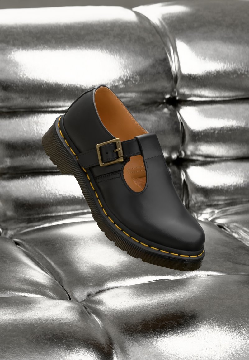 Chaussure Mary Jane en cuir noir avec des surpiqûres jaunes et une boucle en laiton sur une surface matelassée argentée brillante.