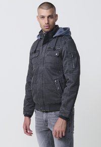 Grå denimjacka med grå huva, flera fickor och ribbade manschetter. Har dragkedjor och tryckknappar; textil med sömnadsdetaljer.