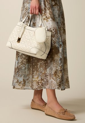 Main tenant un sac à main blanc perforé avec une petite pochette, portant une jupe longue à motifs et des mocassins beige.