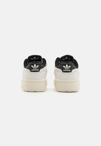 Adidas cipők, főként fehér bőr felsőrésszel, fekete kiegészítőkkel és jellegzetes logóval a sarokrészen. A texturált talp tapadást biztosít.