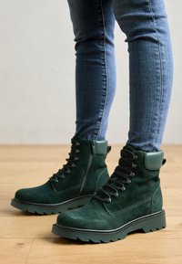 Bottines en daim vert avec une semelle robuste, un avant à lacets et un col rembourré. Fini texturé et fermeture éclair latérale pour un enfilage facile.