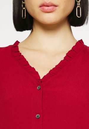 Femme portant un haut rouge à boutons avec un col en V à volants et des boucles d'oreilles en chaîne dorée, montrant uniquement les lèvres et le menton.