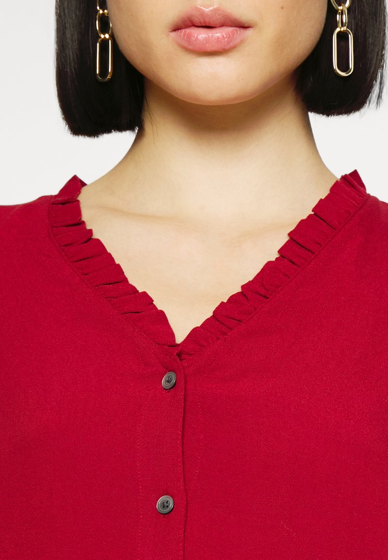 Femme portant un haut rouge à boutons avec un col en V à volants et des boucles d'oreilles en chaîne dorée, montrant uniquement les lèvres et le menton.