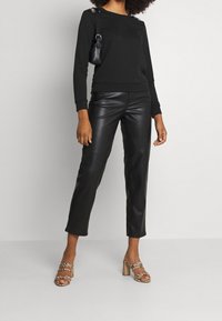 Svart sweatshirt med dekorativa axeldetaljer, matchad med åtsittande svarta läderbyxor och strappar i beige med ett mönstrat band.