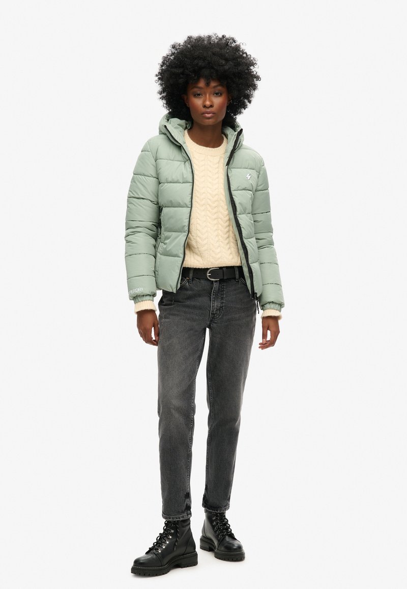 Jasnozielona kurtka puchowa z kapturem, zapinana na zamek, z logo, nałożona na kremowy sweter z dzianiny, w połączeniu z czarnymi dżinsami i botkami.