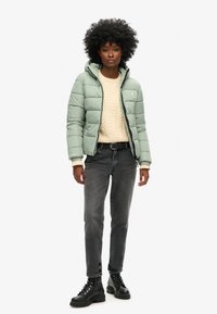 Piumino verde chiaro con cappuccio e design trapuntato. Indossato sopra un maglione lavorato a maglia color crema e abbinato a jeans grigi e stivali neri con lacci.