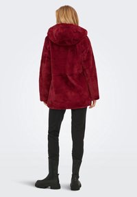 Vrouw die met de rug naar ons toe staat, gekleed in een dieprode nepbont capuchonjas, zwarte slim fit broek en zwarte chunky laarzen.