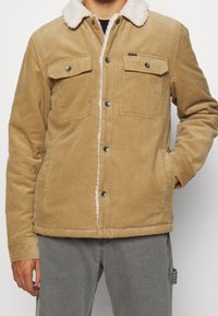 Beżowa kurtka z korduroyu z białym kołnierzem z sztucznego futra, z zatrzaskami i dwoma kieszeniami na piersi. Miękka tekstura z ciepłym, casualowym stylem.