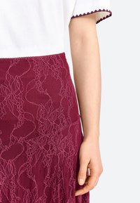 Jupe en dentelle bordeaux avec des motifs floraux, une finition texturée et une taille lisse. Le tissu est semi-transparent avec des détails raffinés.