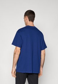 Mand set fra ryggen iført en ensfarvet kongeblå T-shirt med korte ærmer og sorte bukser mod en ensfarvet lys grå baggrund.