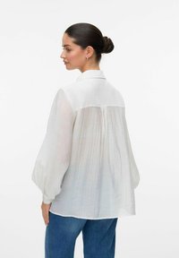Camicia bianca leggera con colletto, schiena plissè e maniche leggermente voluminosi, abbinata a jeans in denim blu.