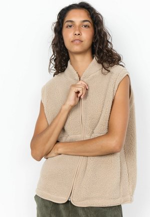Woman with curly hair wearing sleeveless beige fleece vest, holding zipper pull, standing against a white background.
