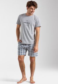 Szara bawełniana koszulka z napisem "HOME RELAX", zestawiona z szortami w niebieską, zieloną i białą kratkę. Bosymi stopami stoi na neutralnym tle.