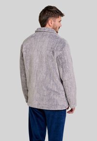 Chaqueta gris con patrón de espiga, con cuello desplegable y corte relajado, combinada con pantalones de terciopelo azul, vista desde la parte posterior.