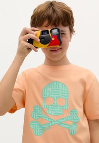 Kompaktní fotoaparát s černým, žlutým a červeným designem. Dítě ho drží blízko. Světle broskvové tričko má design s tyrkysovou lebkou a kostmi.