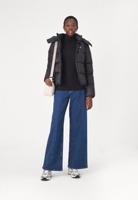 Black puffer jacket with a hood, paired with a black turtleneck and wide-leg blue jeans. Model holds a small cream handbag. Silver sneakers.