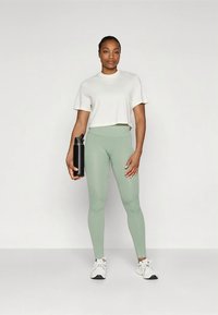 Vaaleanharmaa lyhyt t-paita ja ribbikuvioiset mintunvihreät leggingsit. Musta vesipullo vasemmassa kädessä. Yksivärinen valkoinen tausta. Valkoiset urheilukengät.