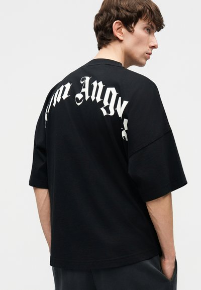 Noor mees lühikeste pruunide juustega, kandes lahtist musta T-särki, mille tagaküljel on osaliselt nähtav valge gooti tekst.