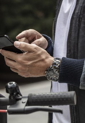 Une personne interagit avec un smartphone, portant une montre noire et argentée. Elle a une manche en tricot bleu et il y a un guidon noir au premier plan.