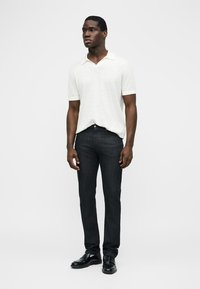 Vyras stovi vilkėdamas baltą trumpomis rankovėmis polo marškinėlį, juodus liesus džinsus ir juodus oficialius batus priešais paprastą foną.